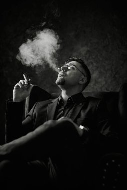 Ciddi bir adamın monokromatik portresi, takım elbise giyiyor ve oturmuş puro içiyor. Duman onu karanlık bir arka planda döndürüyor, sofistike, düşünceli ya da güçlü bir ruh hali yaratıyor.