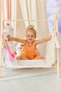 Gülümseyen sarışın bir kız, parlak, rahat bir odada, ahşap bir askıda, kıyafetlerin arasında şakacı bir şekilde oturur. Çocukluk sevincini, bağımsızlığını ve aile hayatını yakalar