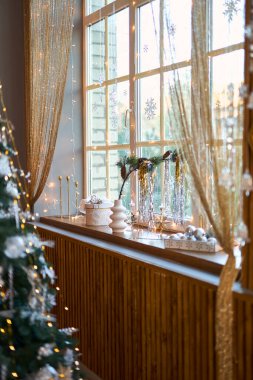 Işıldayan altın renkli perdelerle süslenmiş parlak bir pencere ve berrak kar tanesi süslemeleri, küçük bir Noel dalı ve şenlikli gümüş eşyalar ile pervazda samimi bir tatil sahnesi sergileniyor