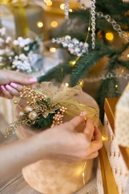 Pembe tırnaklı kadın eller, altın bir yay ve şenlik süslerini güzel bir şekilde sarmalanmış Noel ağacı dalları ve ışıklarıyla çevrili bir hediye kutusuna dikkatlice yerleştiriyor.