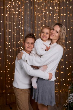 Gülümseyen anne ve beyaz süveter giyen iki çocuğu, sıcacık, şenlikli bir odada, süslenmiş bir ağaç ve altın ışıklarla sarılıyorlar.