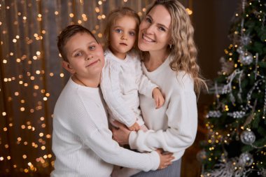 Gülümseyen anne ve beyaz süveter giyen iki çocuğu, sıcacık, şenlikli bir odada, süslenmiş bir ağaç ve altın ışıklarla sarılıyorlar.