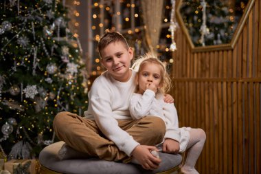 Beyaz süveter giyen bir çocuk ve küçük kız kardeşi süslü bir Noel ağacı ve şenlikli ışıklarla dolu sıcak bir odada birlikte otururken mutlu bir şekilde gülümsüyorlar.