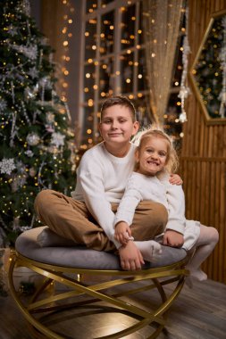 Beyaz süveter giyen bir çocuk ve küçük kız kardeşi süslü bir Noel ağacı ve şenlikli ışıklarla dolu sıcak bir odada birlikte otururken mutlu bir şekilde gülümsüyorlar.