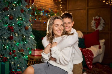 Beyaz süveter giymiş gülümseyen bir anne ve oğlu, Noel ağacı ve ışıklarıyla dolu, rahat, şenlikli bir odada fotoğraf çektirmek için yakın bir şekilde sarılıyorlar.