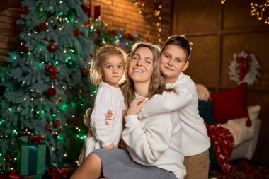 Gülümseyen anne ve beyaz süveter giyen iki çocuğu, sıcacık, şenlikli bir odada, süslenmiş bir ağaç ve altın ışıklarla sarılıyorlar.