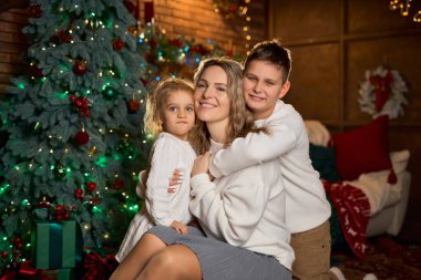 Gülümseyen anne ve beyaz süveter giyen iki çocuğu, sıcacık, şenlikli bir odada, süslenmiş bir ağaç ve altın ışıklarla sarılıyorlar.