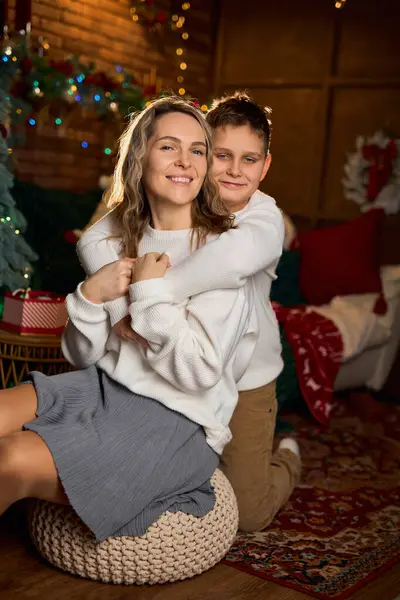 Beyaz süveter giymiş gülümseyen bir anne ve oğlu, Noel ağacı ve ışıklarıyla dolu, rahat, şenlikli bir odada fotoğraf çektirmek için yakın bir şekilde sarılıyorlar.