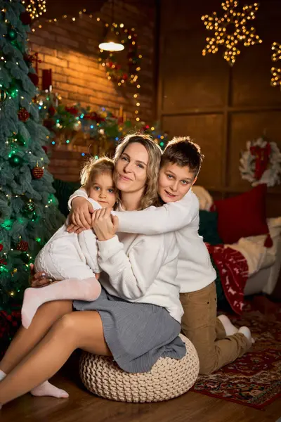 Gülümseyen anne ve beyaz süveter giyen iki çocuğu, sıcacık, şenlikli bir odada, süslenmiş bir ağaç ve altın ışıklarla sarılıyorlar.