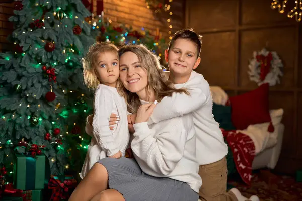 Gülümseyen anne ve beyaz süveter giyen iki çocuğu, sıcacık, şenlikli bir odada, süslenmiş bir ağaç ve altın ışıklarla sarılıyorlar.