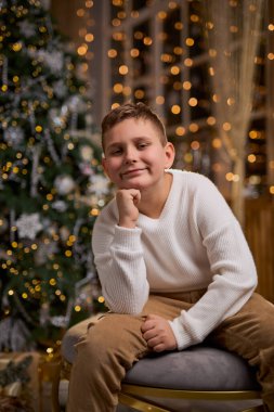 Beyaz kazaklı, gülümseyen bir çocuk, Noel fotoğrafı için düşünceli bir şekilde oturur. Arka planda süslü bir Noel ağacı ve ışıltılı ışıklar vardır.