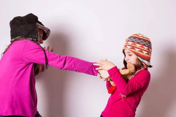 冬の帽子と赤いプルオーバーを身に着けている小さな姉妹は喧嘩し テディベアのために競争します 緑レンガの背景 — ストック写真