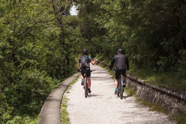 İtalya 'nın Trieste şehrinde Napolyon yolunda bisiklete binen 50' li yaşlardaki arkadaşlar. Kask takıyorlar ve bitkilerle dolu bir patika boyunca bisiklet sürüyorlar. Arkadan bak, dağ bisikletleri üzerinde tanınmayan bisikletçileri göster