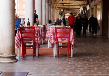 Mantova, Italy - September 15, 2024: Empty tables and chairs outside a restaurant under the city's iconic porticos, with tables featuring the classic Italian red and white checkered tablecloths clipart