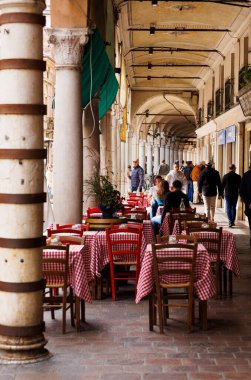 Mantova, İtalya - 15 Eylül 2024: Şehrin ikonik portikolarının altındaki bir restoranın önünde boş masa ve sandalyeler, klasik İtalyan kırmızı ve beyaz kareli masa örtülerinin yer aldığı masalar