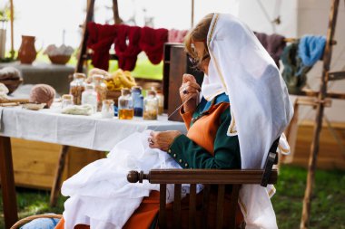 Mantova, İtalya - 15 Eylül 2024: Ortaçağ giysili peçeli bir kadın ahşap bir sandalyede oturur, geleneksel bir dokuma tezgahıyla süslenir, profilde görünür..