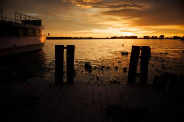Mantua 'daki Mincio Nehri rıhtımında geleneksel ahşap direkler ve kıyıya demir atmış bir tekne bulunuyor. Gün batımında yakalanan sahne, sakin bir siluet etkisi ve sıcak bir atmosfer sergiliyor.