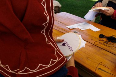 Ortaçağ giysileri içinde peçeli bir kadın ahşap bir sandalyeye oturmuş, geleneksel bir dokuma tezgahıyla nakış işliyor, profilden görülüyor..