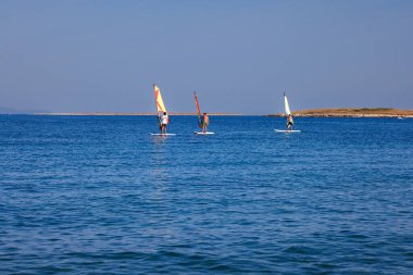 Premantura, Hırvatistan - 22 Ağustos 2024: Hırvatistan 'ın Istria kenti Premantura yakınlarındaki Cape Kamenjak' ta rüzgar sörfü okuluna giden üç erkek. Sahne, kristal berraklığında Adriyatik Denizi 'nde bir açık hava eğitim oturumunu çekiyor