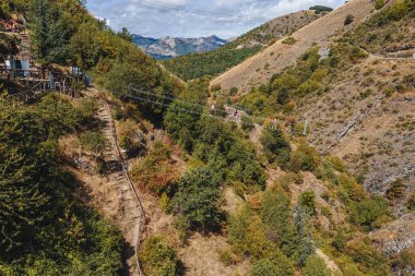 Sasso di Castalda, Italy - August 2024: An aerial view captures a long suspended footbridge   stretching over a rocky terrain with surrounding green and brown foliage, highlighting the adventurous landscape and engineering. clipart