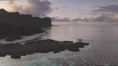 Tropical Maupiti island at sunset, stunning bright sky cloudscape, tranquil sea, mountain silhouette, rugged coastline, small wooden hut on a rocky coast. Remote wild nature scene, travel background clipart