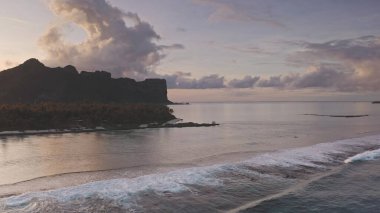 Soft ocean waves gently roll towards silhouette of majestic mount Teurafaatiu, Maupiti island at pink sunset serene breathtaking scene. Remote wild nature paradise, exotic summer travel. Aerial flight clipart