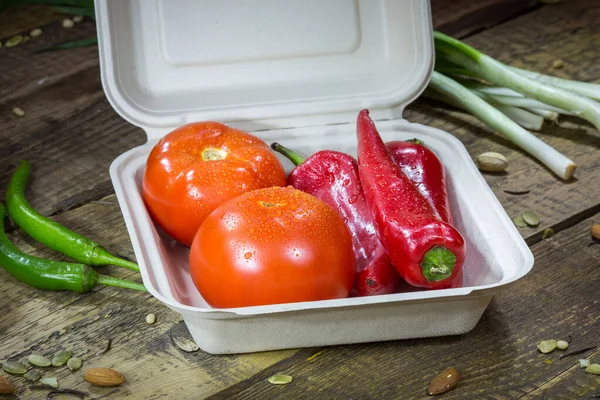 Paprika, domates ve yeşillikler ahşap bir arka planda tek kullanımlık bir kabın içinde.