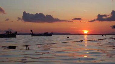 Güneş sakin bir körfezin üzerinde batarken gökyüzü turuncu parlıyor, denizden yansıyor. Tekneler kıyıya sürüklenirken martılar uzak bir ufukta belirdi.