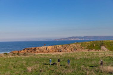 La Coruna, İspanya: A Coruna sahilinde güneşli bir kış gününün tadını çıkaran insanlar, açık mavi bir gökyüzü ve denizin ötesindeki uzak tepeler