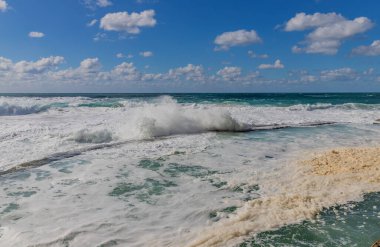 Dalgalar, Azenhas do Mar 'daki kayalıklara çarpıyor. Mavi gökyüzünün altında deniz köpüğü uçuşuyor..