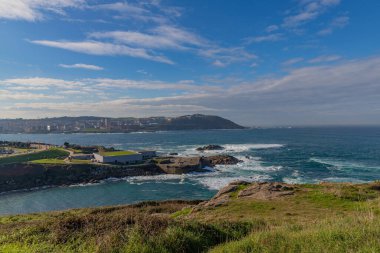 A Coruna 'da kıyı manzarası. Çimenli arazi, kayalık kayalıklar ve kayalara çarpan dalgalar.