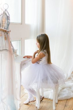 young beautiful ballet ballerina sits on an ottoman in front of a mirror. Little girl in a white dress.