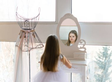 young beautiful ballet ballerina sits on an ottoman in front of a mirror. Little girl in a white dress.
