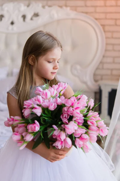 child gives flowers. portrait of a little girl with a bouquet of pink tulips flowers. a girl in a white dress. Bright interior.