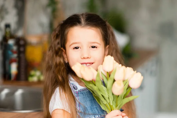 Smiling child gives flowers. portrait of a little girl with a bouquet of tulips flowers. girl in a denim dress. Gray interior. Mother's Day, birthday.