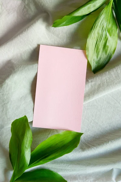 亚麻布桌布背景上的空白问候 粉红卡片和绿色大叶子 平躺在地上 尽收眼底 夏天的婚礼 母亲节文具的模拟场景 垂直照片 — 图库照片