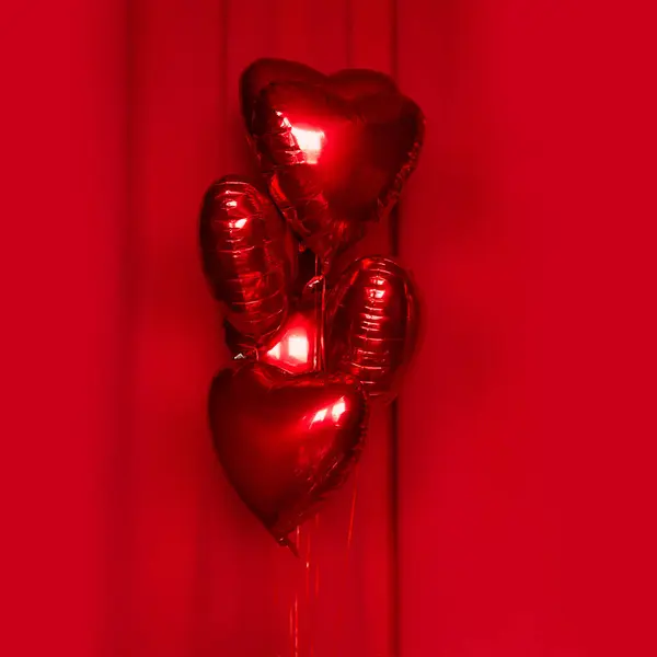 Red metallic helium balloons in the shape of a heart on a red fabric background. Foil balloons. Minimal love concept. Decoration for Valentine\'s Day or wedding party.