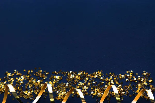 Confetti stars and gold streamer on dark blue background, flat lay. Christmas celebration card, blank mockup.