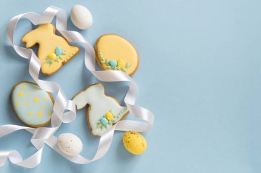 Paskalya 'da ev yapımı zencefilli çörek ya da kremalı kurabiye, tavşan ve yumurta desenli. Beyaz çikolatalar, paskalya yumurtaları. Beyaz kurdeleli dekor. Mavi arkaplan, üst görünüm, kopyalama alanı