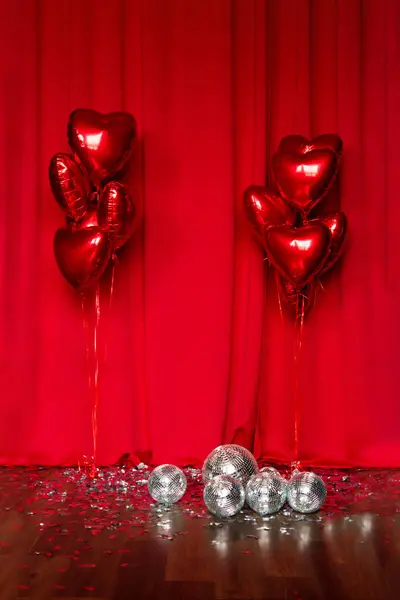 Red metallic helium balloons in the shape of a heart on a red fabric background. Foil balloons. Minimal love concept. Decoration for Valentine's Day or wedding party. Silver disco balls on the floor.