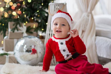 Noel Baba kostümü ve kırmızı şapkalı gülümseyen küçük bebek bir Noel ağacının önünde yumuşak bir kürk halısında oturuyor.