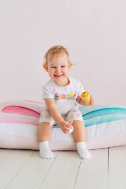 Doğal kumaştan yapılmış beyaz bir tişörtle küçük, sevimli, sağlıklı bir kız çocuğu, kameraya bakıyor, oyuncaklarla oynuyor, yastığa oturuyor, bebek evde.