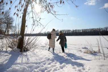 İki kadın kız arkadaş kış parkında karlı manzaraların önünde yürüyor.