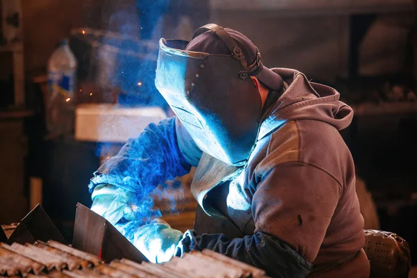 Factory worker in protective mask welding iron parts.
