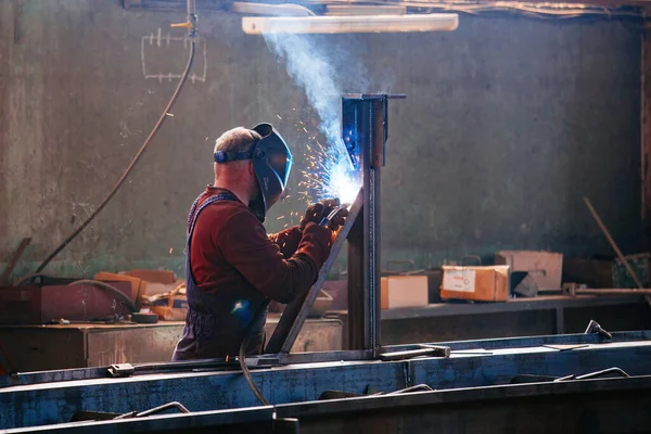 Factory worker in protective mask welding iron parts.