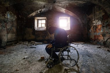 Terk edilmiş hastane koğuşunda eski tekerlekli sandalyede oturan üzgün adam. Eski binadaki engelli hastalar için bozulmuş araçtaki siluet.