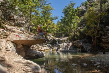 Young good looking woman summery dressed, wearing white beach hat, sitting at the small cliff rock over the water, surrounded by deep green forest, environmental friendly 