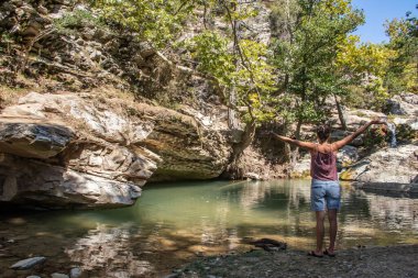 Mavi şort, pembe gömlek ve beyaz yaz şapkası giymiş genç bir kadın, küçük bir orman gölünde duruyor güzel bir doğanın ve temiz havanın keyfini çıkarıyor.
