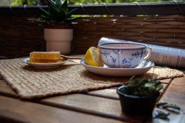 Balkondaki ahşap masa, okunacak kitaplar, bir fincan doğal çay, çaydanlık, çiftlikten gelen organik bal, taze yeşil çay yaprakları ve organik meyveler, arka planda binalar arasındaki küçük park manzarası, dinlenmek için küçük vaha.