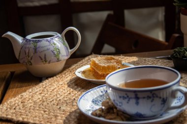 Balkondaki ahşap masa, okunacak kitaplar, bir fincan doğal çay, çaydanlık, çiftlikten gelen organik bal, taze yeşil çay yaprakları ve organik meyveler, arka planda binalar arasındaki küçük park manzarası, dinlenmek için küçük vaha.